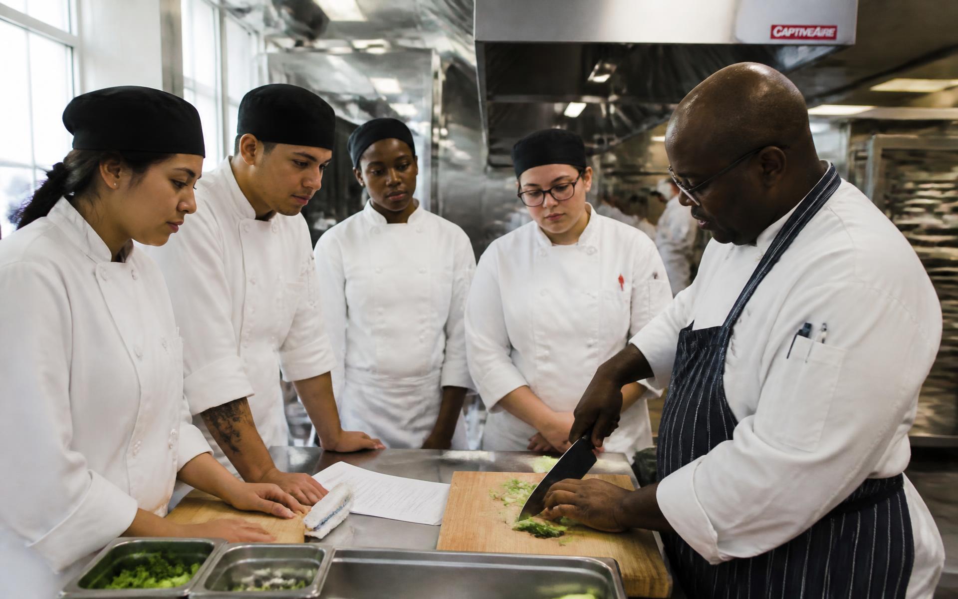 Culinary trainees learning knife skills from a chef instructor inside a professional commercial kitchen